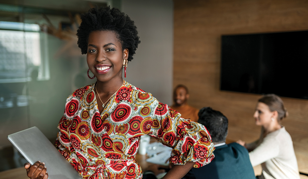 Beautiful young smiling professional black african business woman holding laptop, coworkers hold a meeting in background